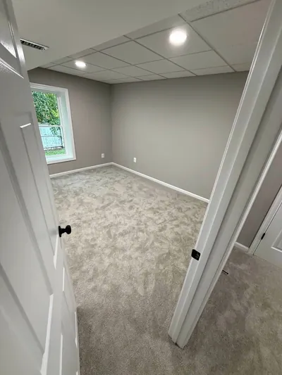 A finished basement bedroom seen through an open door: gray carpet, a window and recessed lights in a drop ceiling
