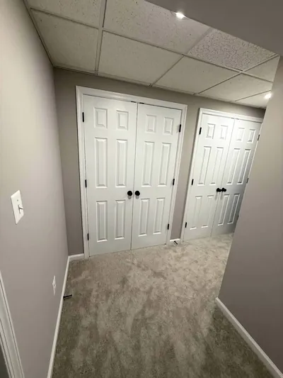 A carpeted basement room with two sets of white double closet doors under a drop ceiling