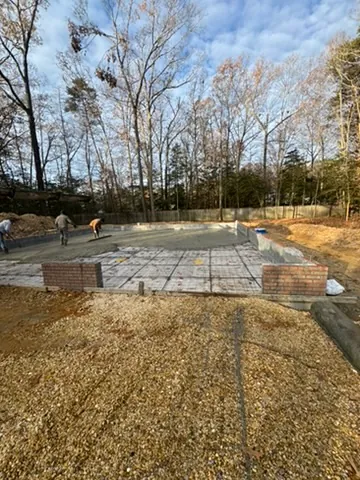 A block foundation with brick piers and a prepared slab on a wooded lot, gravel in the foreground and the crew at work in the distance