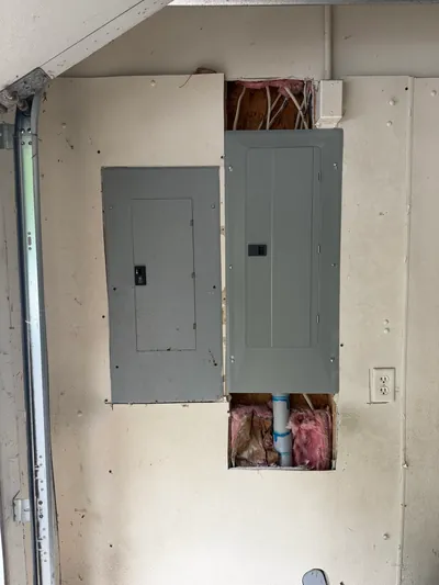 Two gray electrical panels set side by side in a garage wall, the drywall opened above and below them for new cable