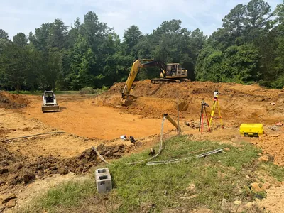 An excavator and a skid steer cutting a foundation into red clay, with survey tripods set up at the edge of the dig
