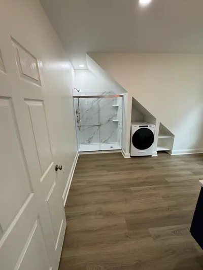 A glass-door shower with a marble-look surround beside a front-loading washer, tucked under the roof slope
