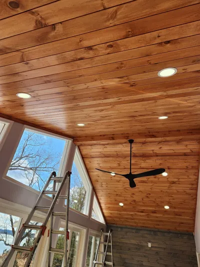 A vaulted knotty pine ceiling with recessed lights and a black ceiling fan, ladders still standing below