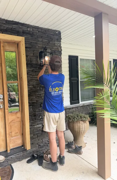 A crew member in a blue Stamp Electric shirt, seen from behind, fitting a lantern light to a stacked stone wall beside a wood front door