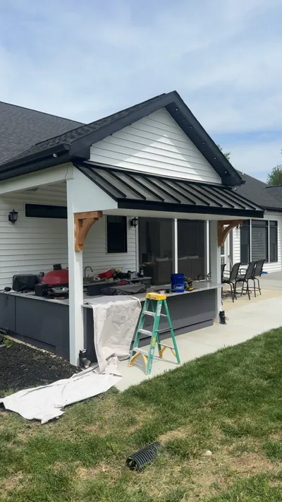 The back of a white-sided house with a covered porch under a black standing seam metal roof on wood brackets, an outdoor kitchen counter beneath it and drop cloths still on the lawn