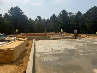 A new concrete slab inside its foundation walls, framing lumber stacked beside it and the crew at work on the far side, tall pines behind